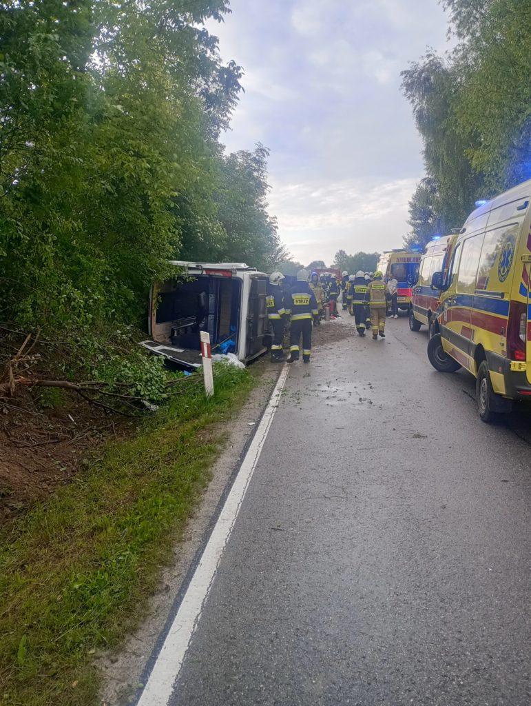 Autobus przerwrócony na bok, obok strażący w strojach bojowych, trzy karetki po prawej stronie