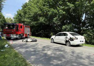 Rozbity motocykl leży na drodze, obok czerwony samochód strażaków, na pierwszym planie białe auto osobowe