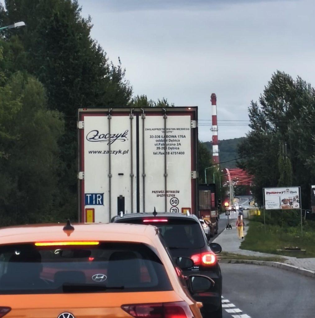 Samochody stoj ace w korku, drzewa obok drogi, na horyzoncie komin fabryczny