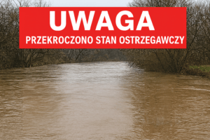 Wezbrana rzeka z mętną wodą, u góry czerwone pole z napisem uwaga