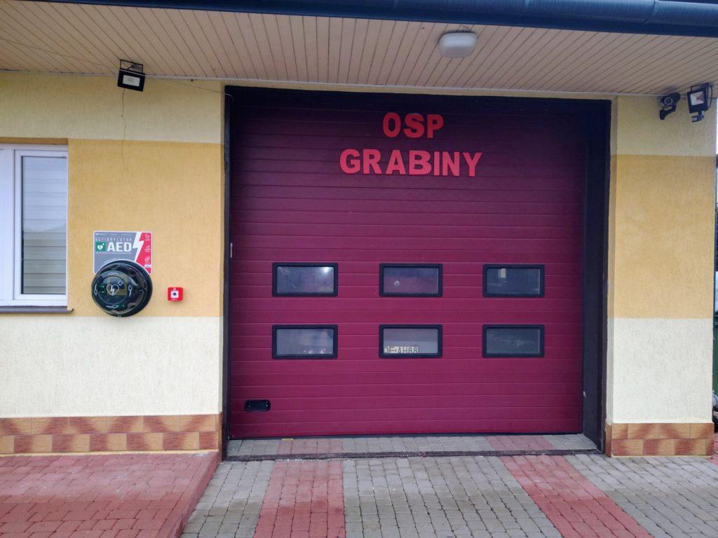 drzwi garażowe z napisem OSP Grabiny obok defibrylator