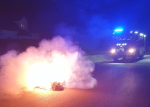 Zdjęcie przedstawia nocną akcję straży pożarnej przy pożarze balotu słomy. Na pierwszym planie widoczny jest płonący balot, z którego wydobywa się intensywny ogień i gęsty dym. W tle znajduje się wóz strażacki z włączonymi niebieskimi sygnałami świetlnymi, oświetlającymi drogę i miejsce zdarzenia. Fotografia oddaje dramatyzm interwencji oraz skalę zagrożenia, z jakim musieli zmierzyć się strażacy.