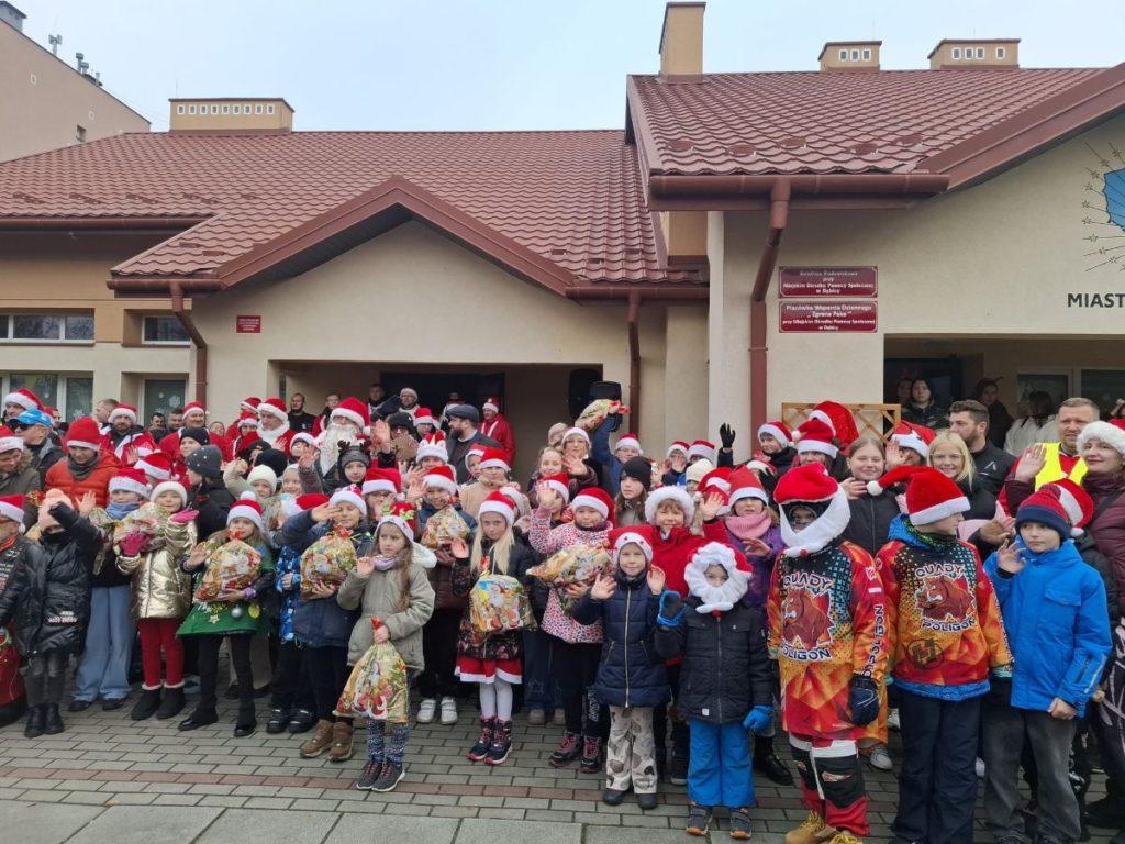 Grupa dzieci i dorosłych w czapkach Mikołaja pozujących przed budynkiem placówek „Zgrana Paka” w Dębicy podczas wizyty Motomikołajów. Uśmiechnięte dzieci trzymają świąteczne paczki, a obok nich stoją motocykliści w kolorowych strojach, tworząc radosną i pełną energii przedświąteczną atmosferę.