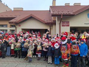 Grupa dzieci i dorosłych w czapkach Mikołaja pozujących przed budynkiem placówek „Zgrana Paka” w Dębicy podczas wizyty Motomikołajów. Uśmiechnięte dzieci trzymają świąteczne paczki, a obok nich stoją motocykliści w kolorowych strojach, tworząc radosną i pełną energii przedświąteczną atmosferę.