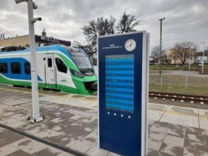 Na pierwszym planie niebieska talica informacyjn, za nią jet pociąg w zielono-niebiesko-białym kolorze