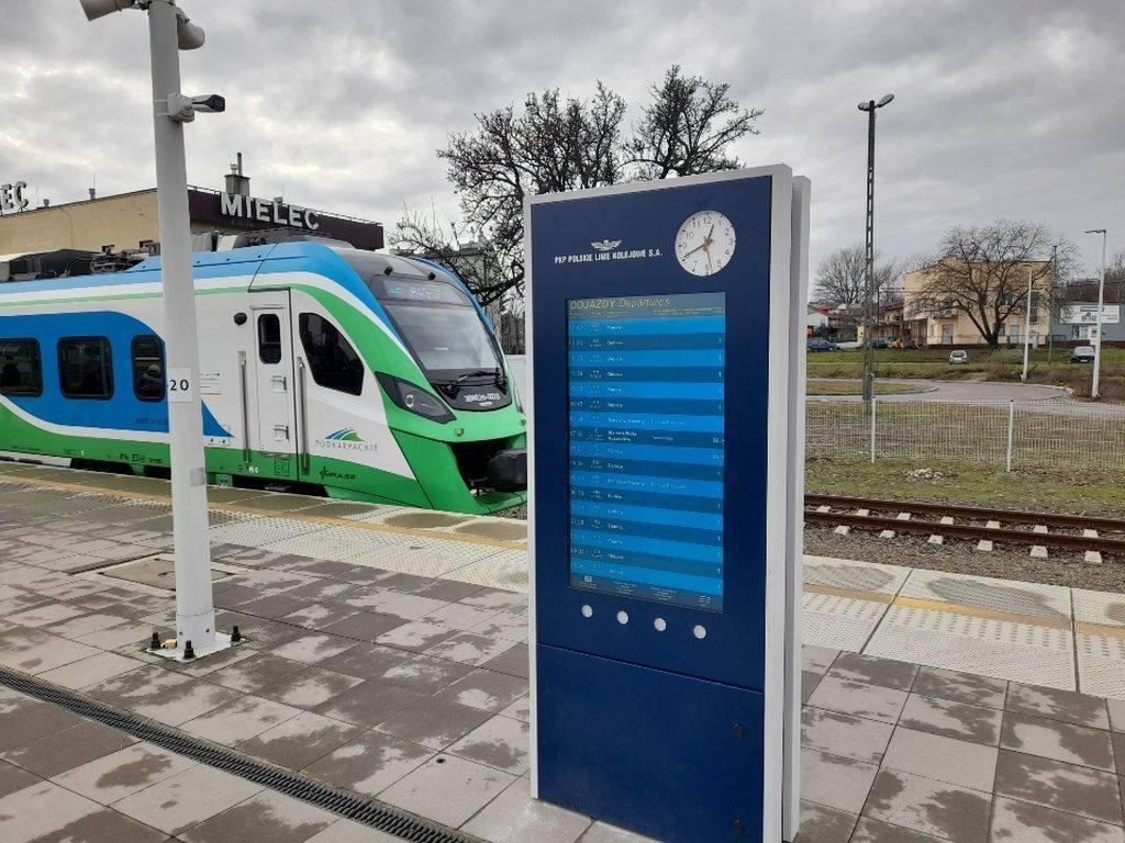 Na pierwszym planie niebieska talica informacyjn, za nią jet pociąg w zielono-niebiesko-białym kolorze