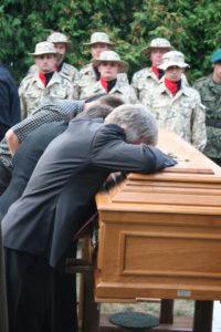 drewniania trumna z ciałem żołnierza, nad nią pochylone trzy osoby w żałobie, w tle żołnierze w mundurach i czarno-czerwonych chystach pod szyją