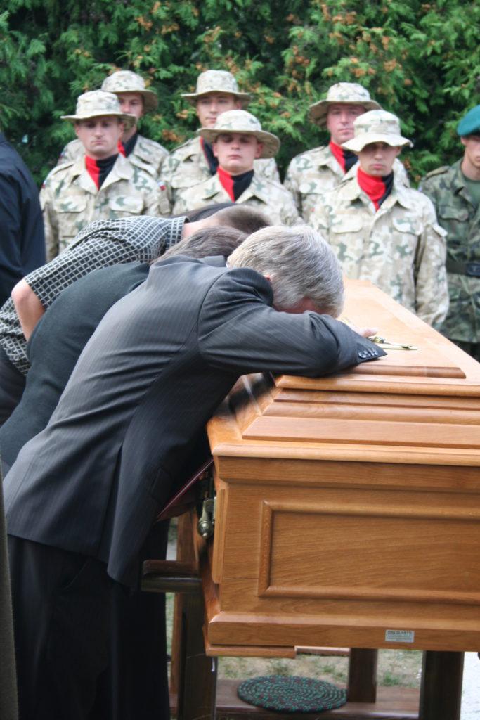 drewniania trumna z ciałem żołnierza, nad nią pochylone trzy osoby w żałobie, w tle żołnierze w mundurach i czarno-czerwonych chystach pod szyją