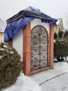murowana kapliczka o białych i ceglanych ścianach, przykryta folią niebieską i czarną