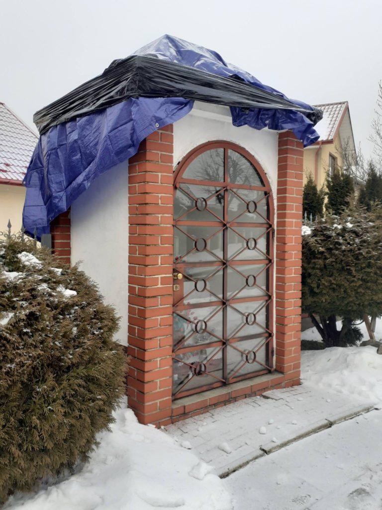 murowana kapliczka o białych i ceglanych ścianach, przykryta folią niebieską i czarną