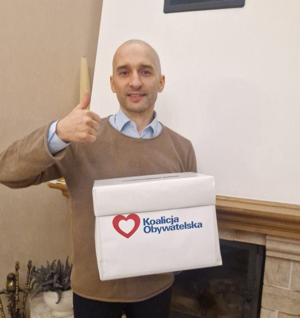 Młódy mężczyzna , uniesiony kciuk w górę, w ręku trzyma plakat z napisem Koalicja Obywatelska