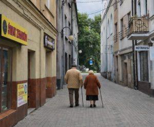 Dwoje starszych ludzi idących wąską uliczką w centrum miasta, podpierających się laskami.