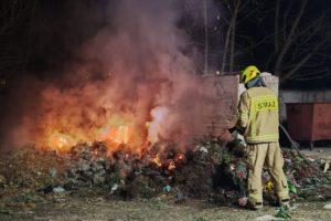 Paląca się sterta śmieci, strażak w stroju bojowym gasi ogień.